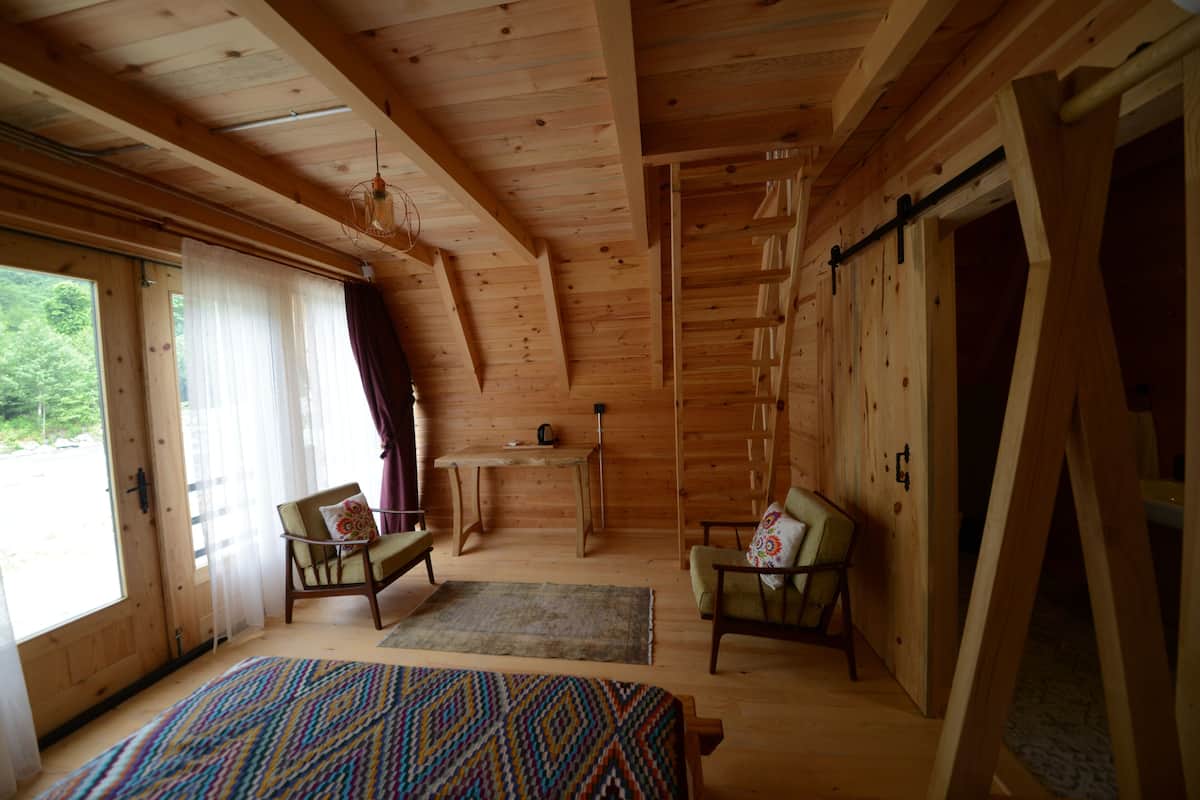 Una habitación de cabaña de madera con una cama, dos sillas, una mesa y un ventanal con cortinas.