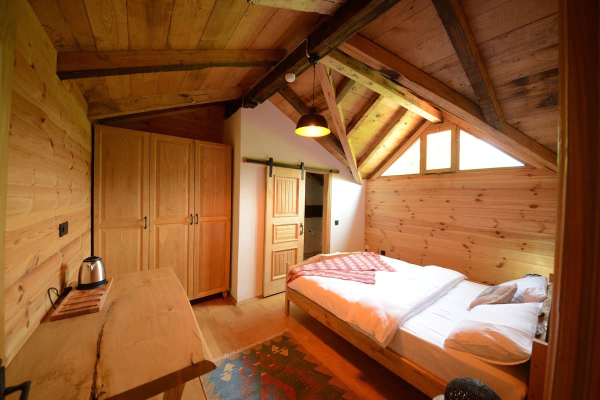 Un dormitorio de cabaña de madera con techo alto, una cama, una mesa con hervidor y una puerta que da a otra habitación.