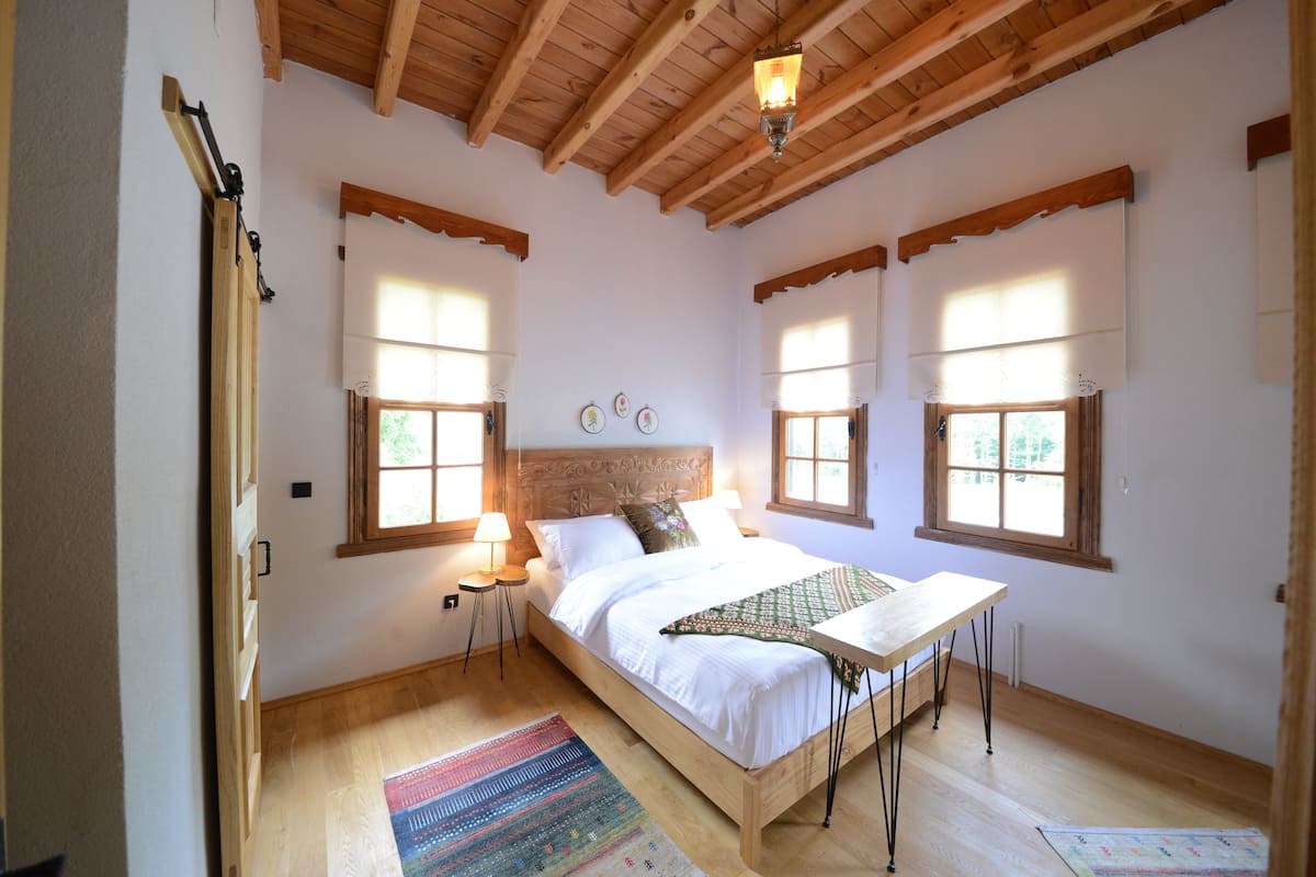 Un dormitorio con una cama de madera, una mesita de noche, dos lámparas de pared y una ventana con vistas a la vegetación.