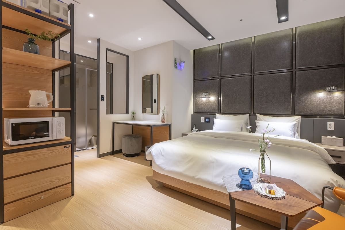 A modern hotel room with a large bed, a wooden desk, a microwave, and a bedside table with a vase.