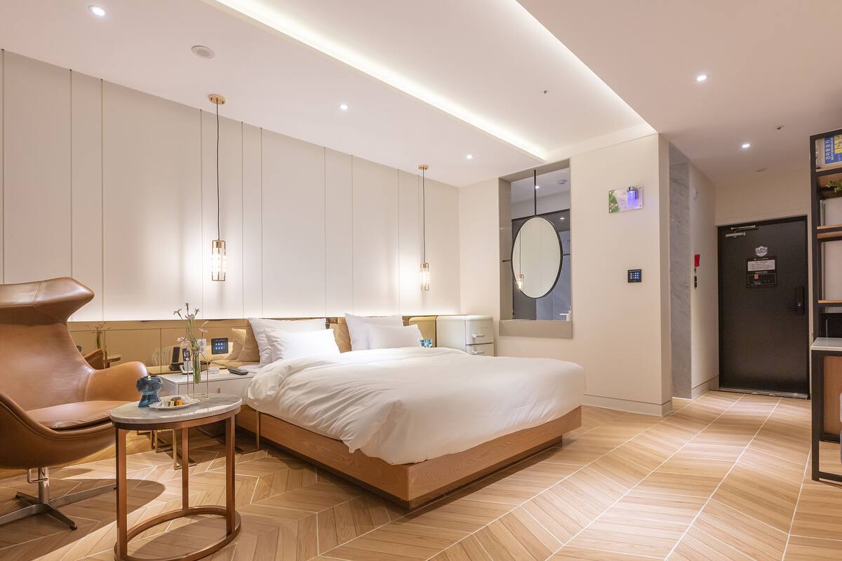 A modern hotel room with a large bed, a sitting area with a chair and table, a bedside table with a lamp, and a large mirror on the wall.