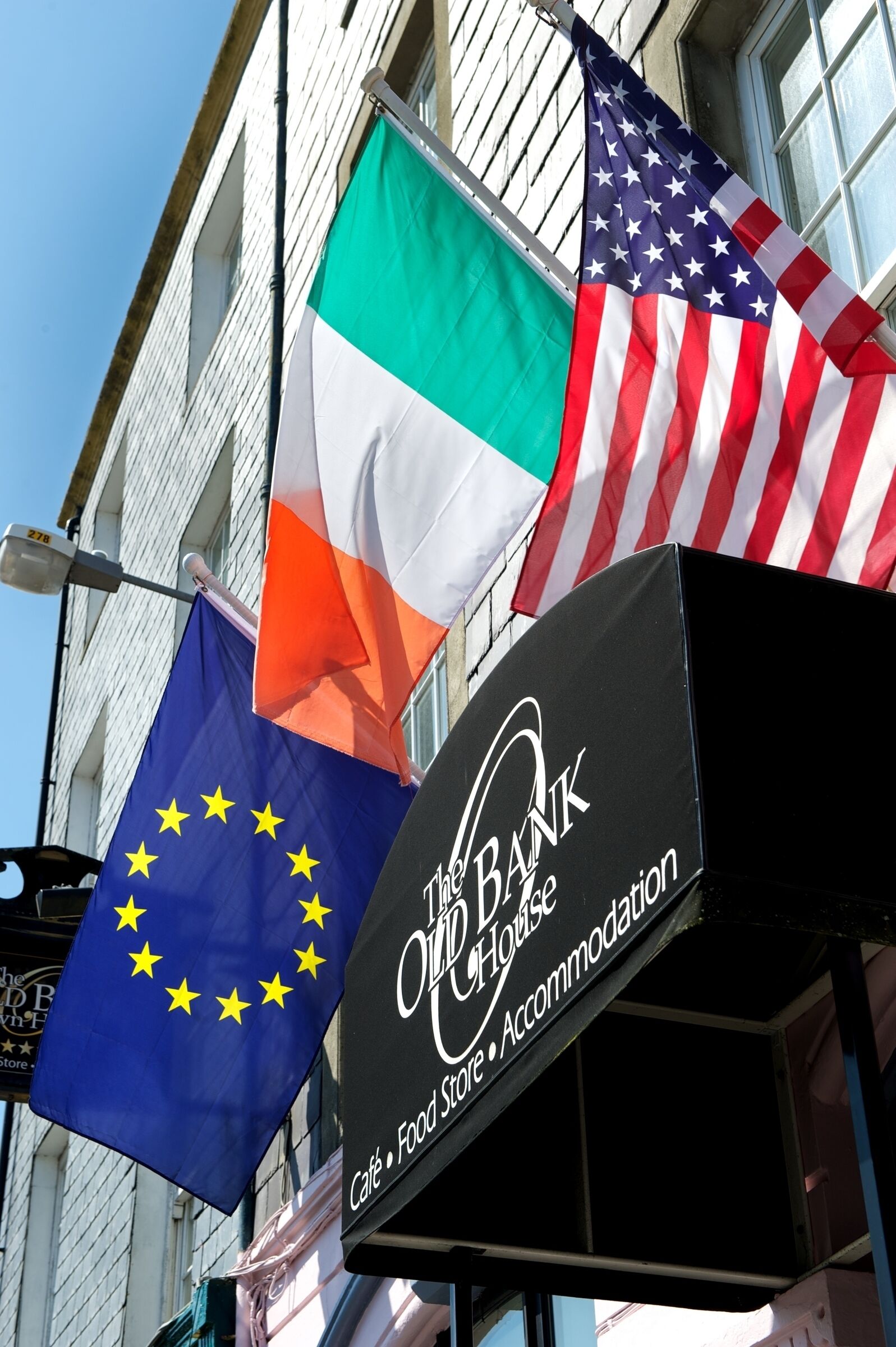 A building with multiple flags, including the EU, US, and Irish flags, and a sign for 'The Old Bank House Café, Food Store, Accommodation'.