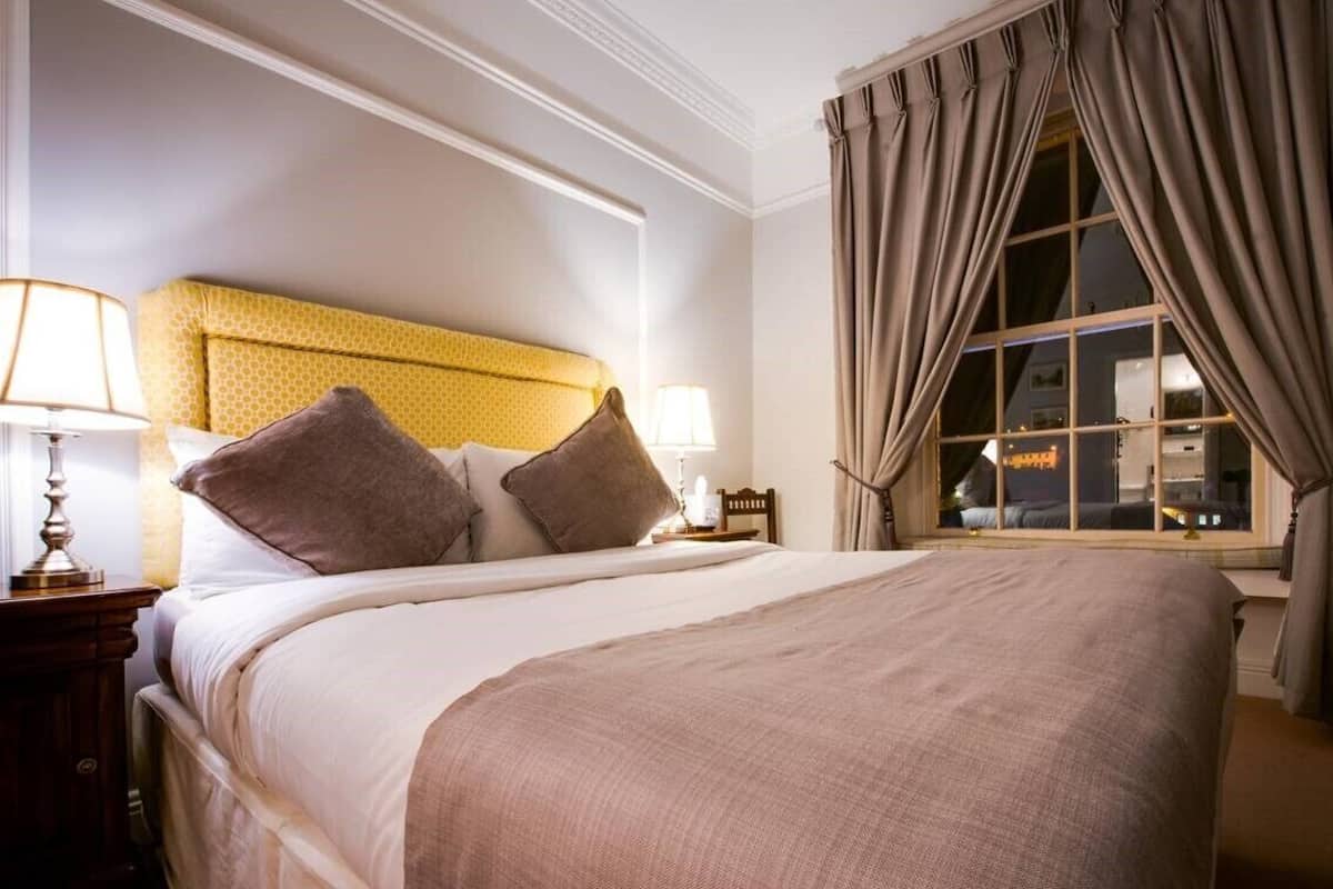 A neatly made bed with a headboard, two bedside lamps, and a window with curtains.