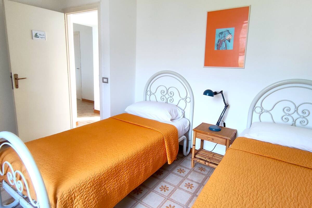 A room with two beds, a nightstand with a lamp, a door, and a wall with a framed picture.