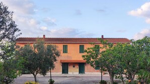 A building with a red-tiled roof and green shutters, surrounded by trees and yellow flowers. - Eco House San Michele (Latina)