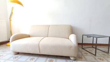 A beige sofa with a wooden base, a small side table, and a yellow floor lamp in a room with patterned floor tiles.