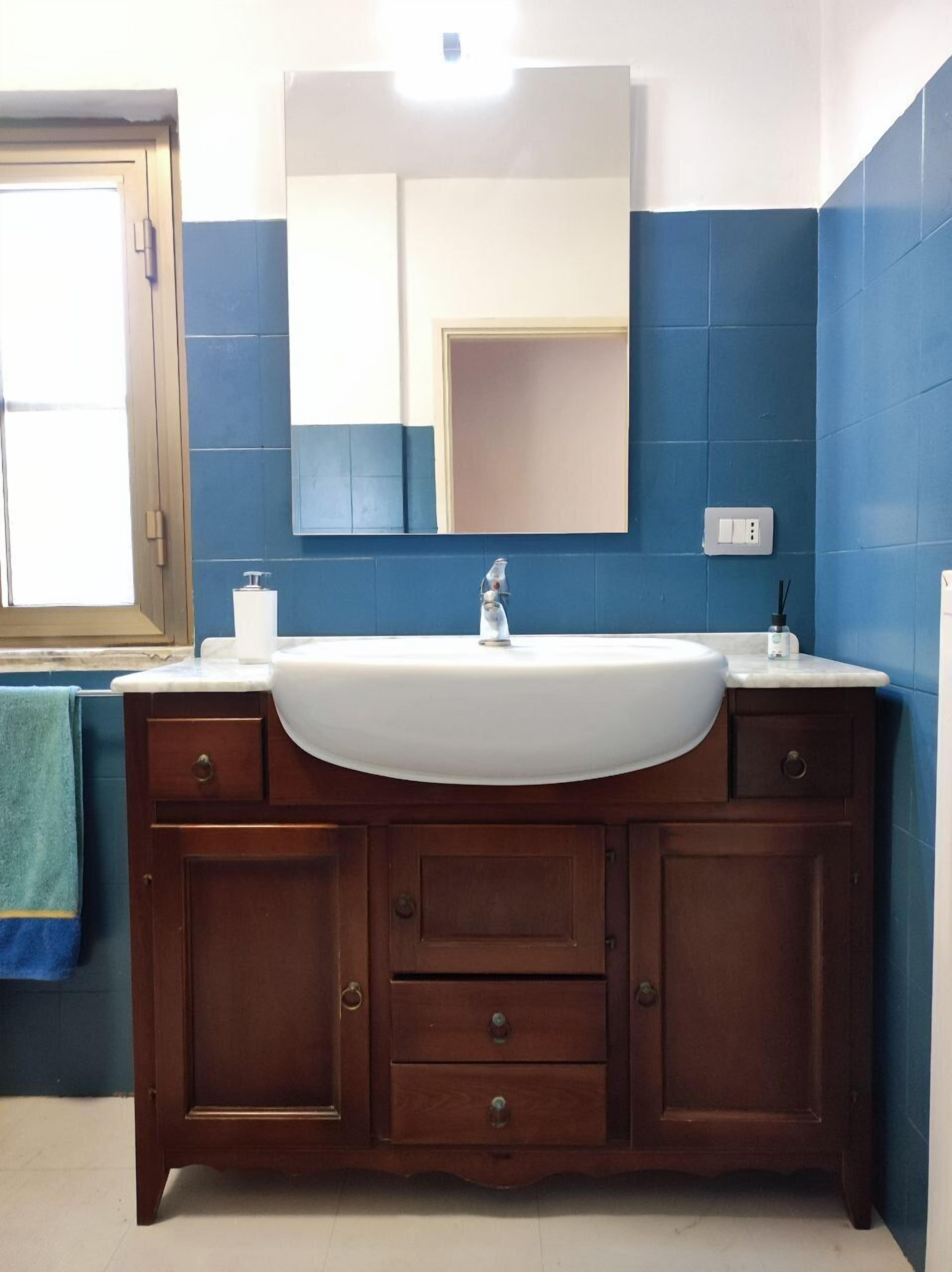 Een badkamer met een wit wastafelmeubel, houten kast, blauwe tegels, een spiegel en een handdoekhouder.
