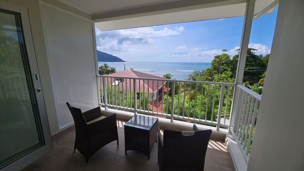 A balcony with two wicker chairs and a small table, overlooking a coastal view with houses and greenery. - Modern and Private - 1min from the Beach! (Beau Vallon)