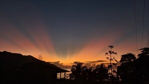 A sunset over a coastal landscape with silhouettes of mountains, trees, and a small structure. - Modern and Private - 1min from the Beach! (Beau Vallon)