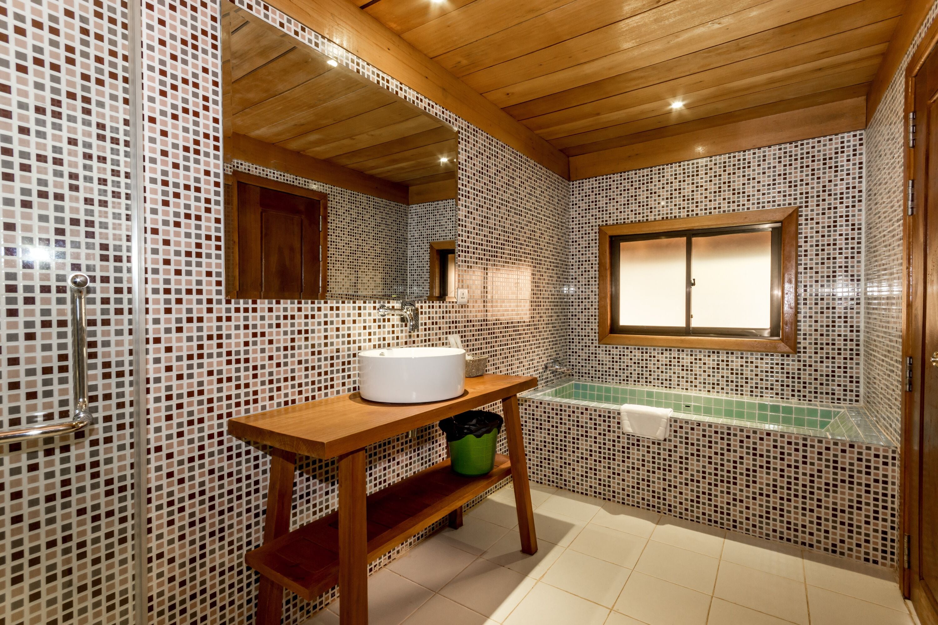 Une salle de bain aux murs recouverts de mosaïques, un meuble-lavabo en bois avec un évier, une poubelle verte et une fenêtre sur laquelle est accrochée une serviette.