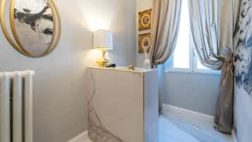 A bathroom with a marble vanity, a round mirror with a gold frame, a lit lamp, and a window with curtains.