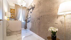 A hallway with marble flooring, a round gold table with a vase of white flowers, a large wall mural, and a chandelier. - Le Dimore Suites Milano (Milan)