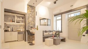 A modern living room with a grey sofa, a coffee table, and a dining area with a kitchen counter. - CAPRI Downtown Rooms (Mykonos)
