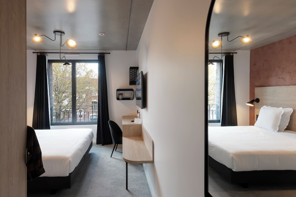 Une chambre d’hôtel avec deux lits simples, un bureau, une chaise, une télévision et une grande fenêtre avec des rideaux.