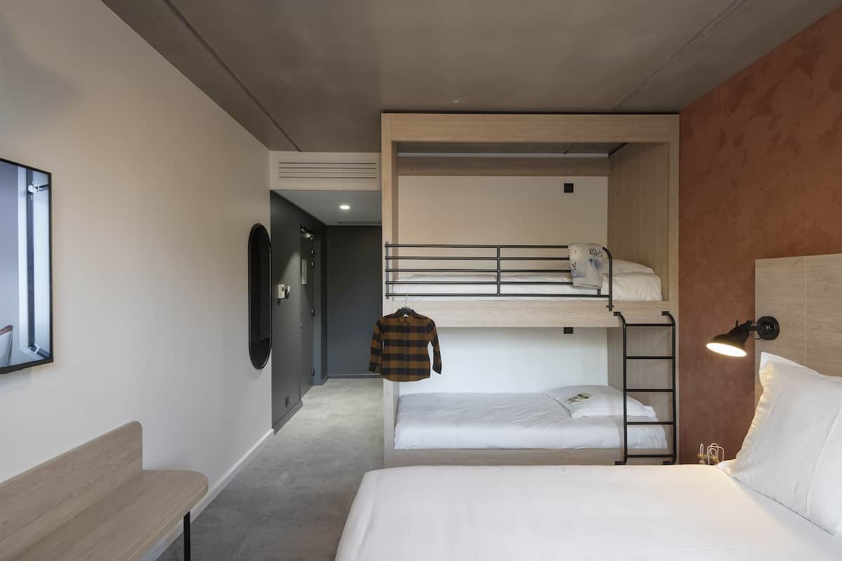 A room with a bunk bed, a single bed, a bench, a wall-mounted TV, and a bedside lamp.