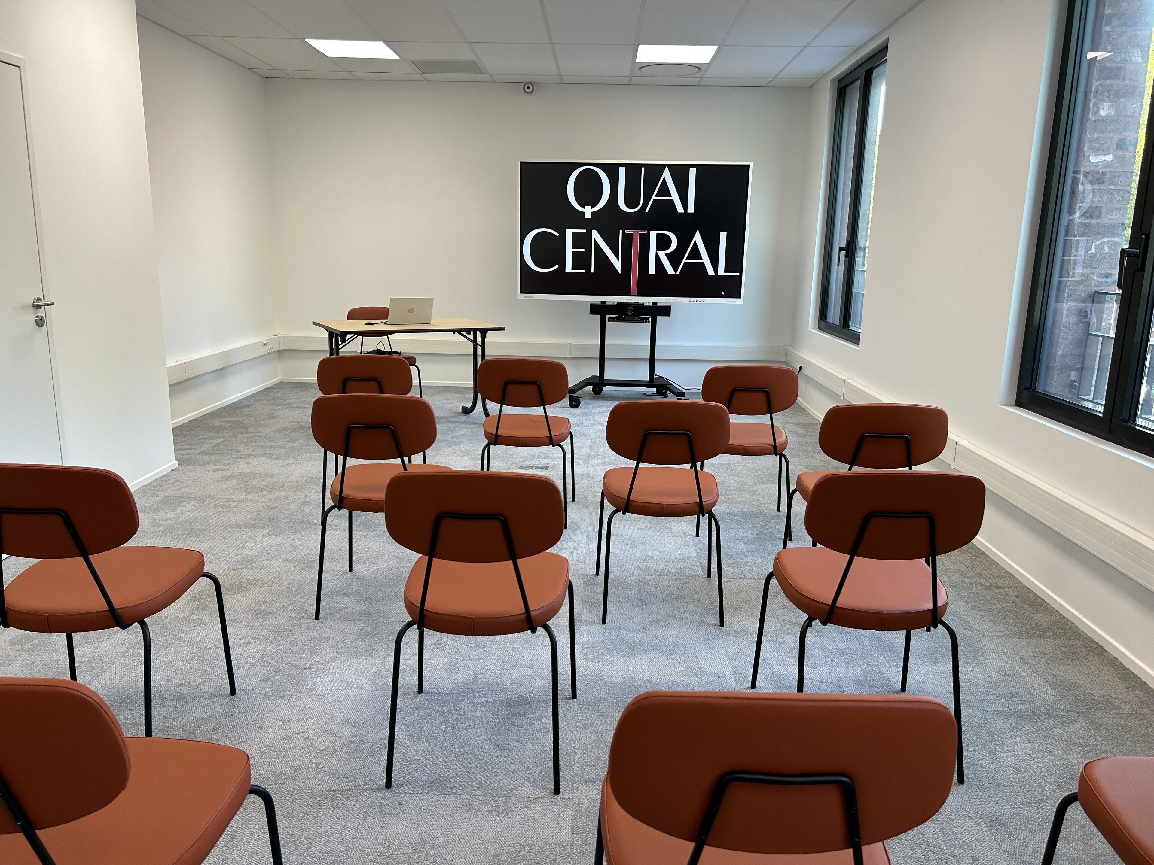 A conference room with a large screen displaying "QUAI CENTRAL" and several orange chairs arranged around a table with a laptop.
