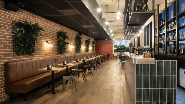 A restaurant with long wooden tables, cushioned booths, and a brick wall with greenery. - Quai Central (Tourcoing)