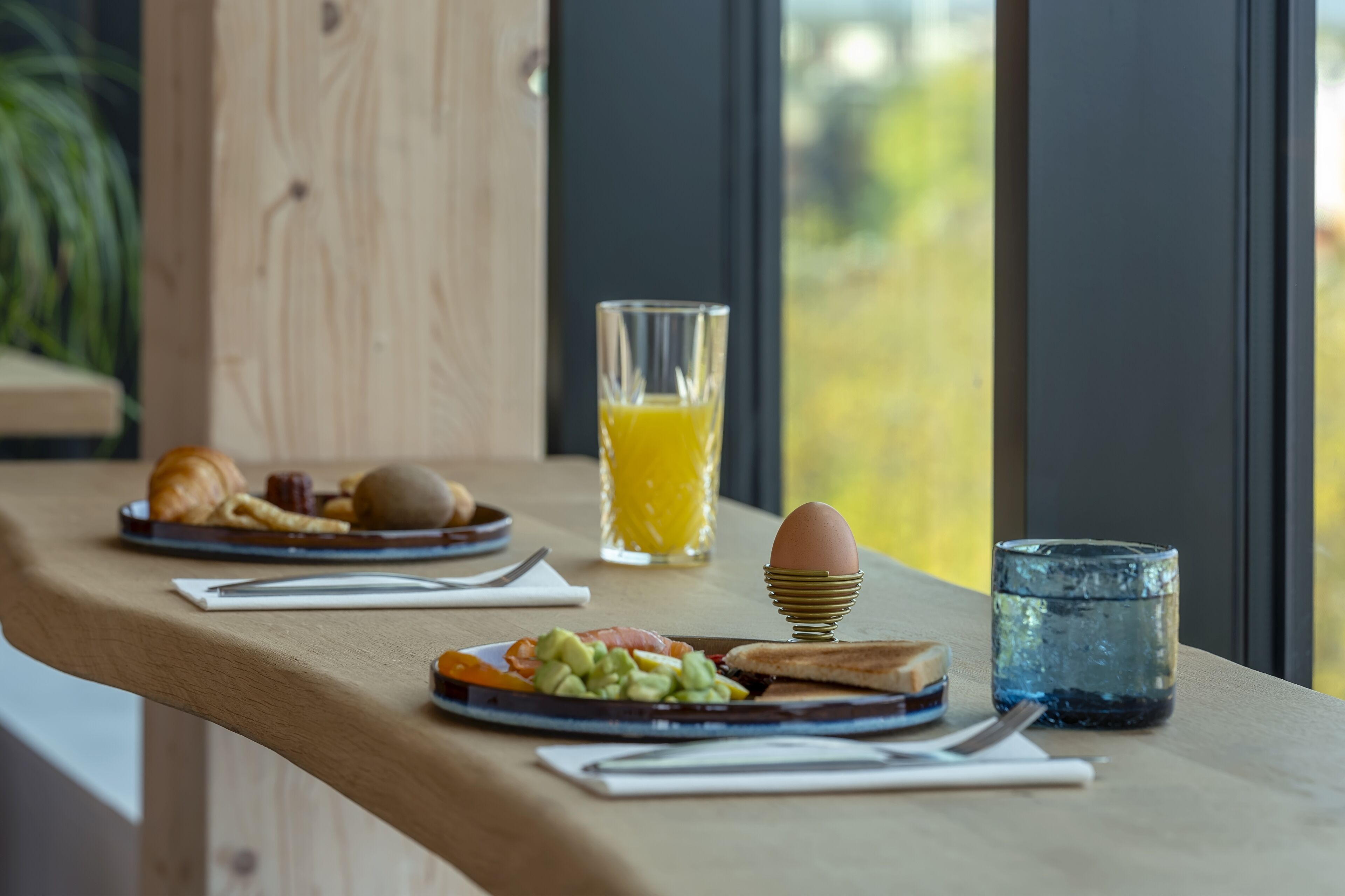 Una mesa de desayuno con cruasanes, un plato de huevos, aguacate y tostadas, un vaso de zumo de naranja y un vaso de agua.
