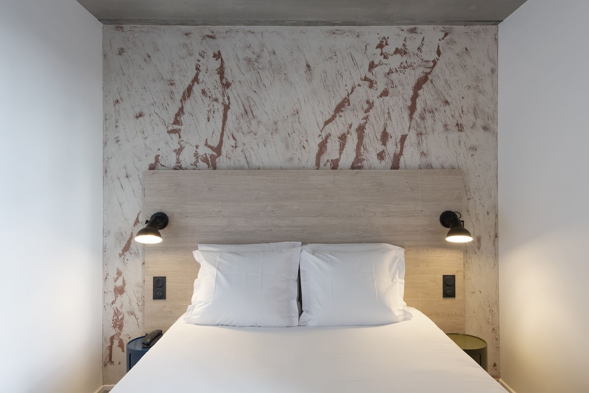 A hotel room with a wooden headboard, two bedside lamps, a bed with white linens, and a small bedside table with a lamp.