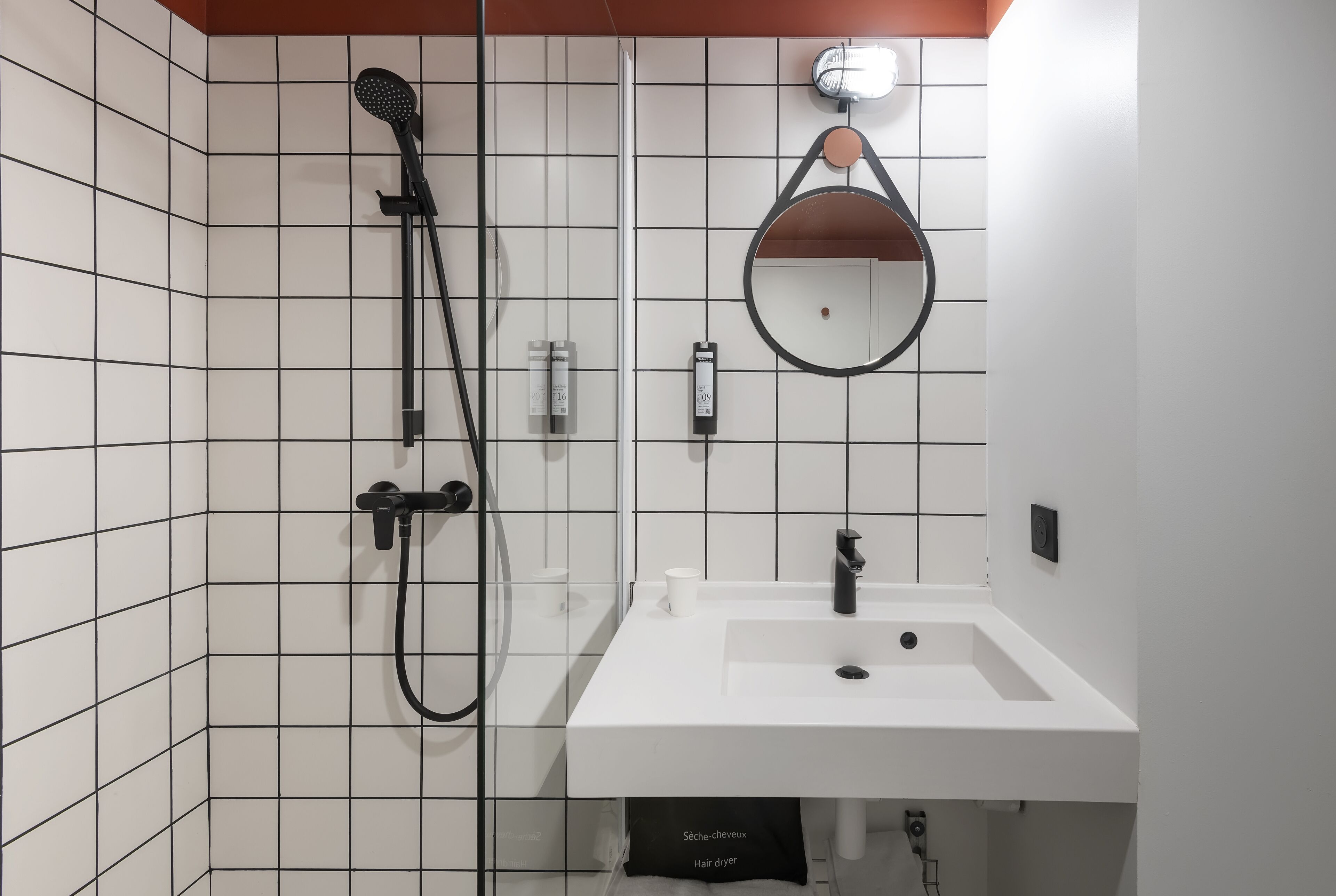A bathroom with white tiled walls, a white sink, a black handheld showerhead, a round mirror, and a hair dryer.