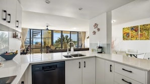 A modern kitchen with white cabinets, a dishwasher, and a large window offering a view of palm trees. - Beachfront, ocean views, private rooftop terrace with spa, pool, walk to beach (Marcoola)