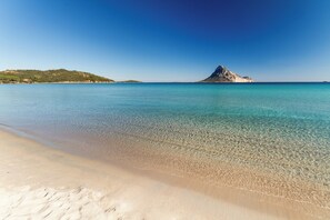 A beach with clear turquoise water, a sandy shore, and a rocky hill in the distance. - Stunning view near the beach with large panoramic veranda MOLARA house (Porto Taverna)