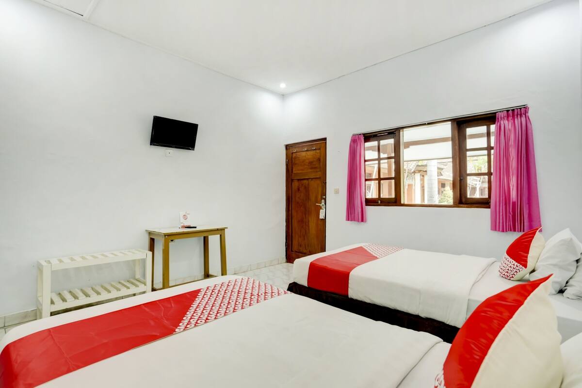 Habitación de hotel con dos camas, una televisión, una puerta de madera, una ventana con cortinas rosadas y una mesita pequeña.