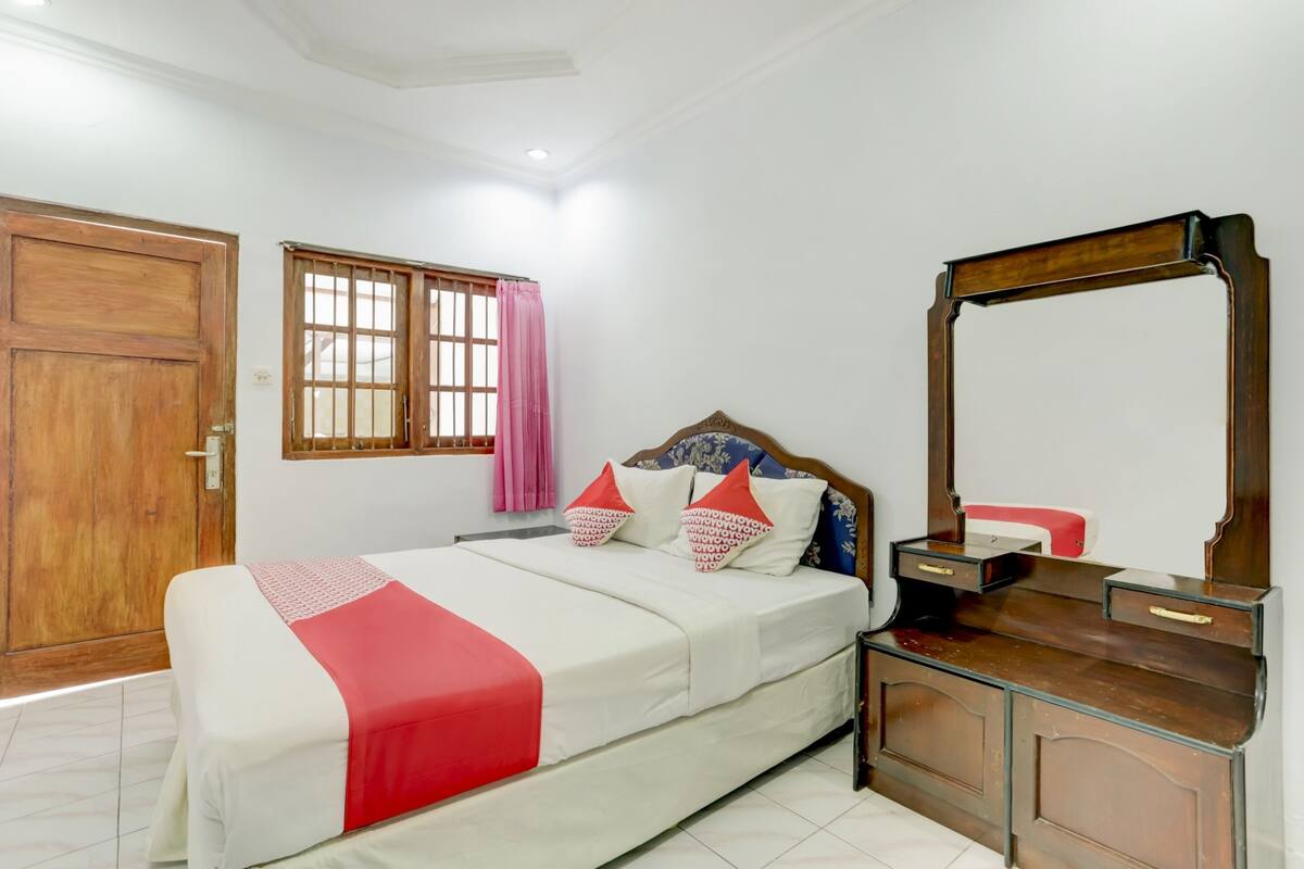 Un dormitorio con puerta de madera, una ventana con cortinas, una cama con colcha roja y blanca, y un aparador de madera con espejo.