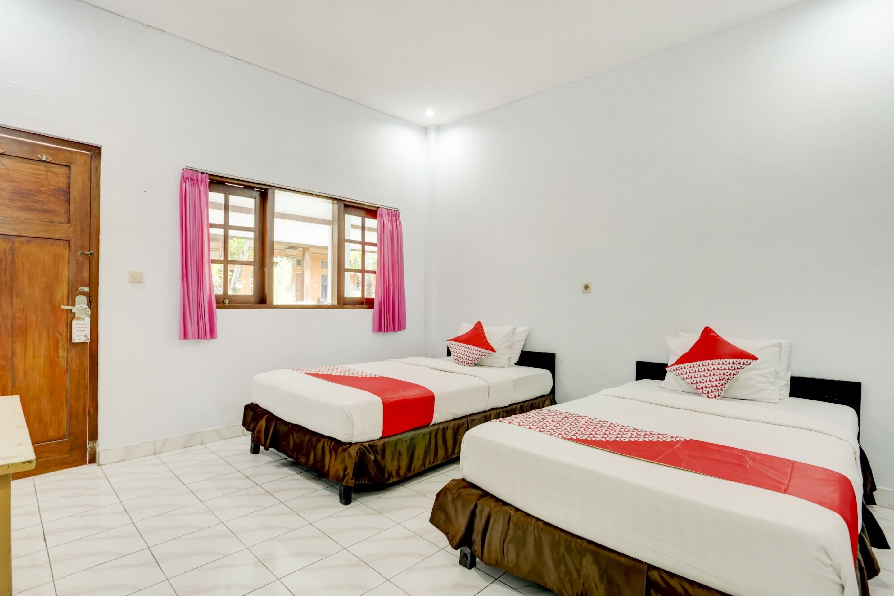 A room with two beds, a wooden door, a window with pink curtains, and a tiled floor.