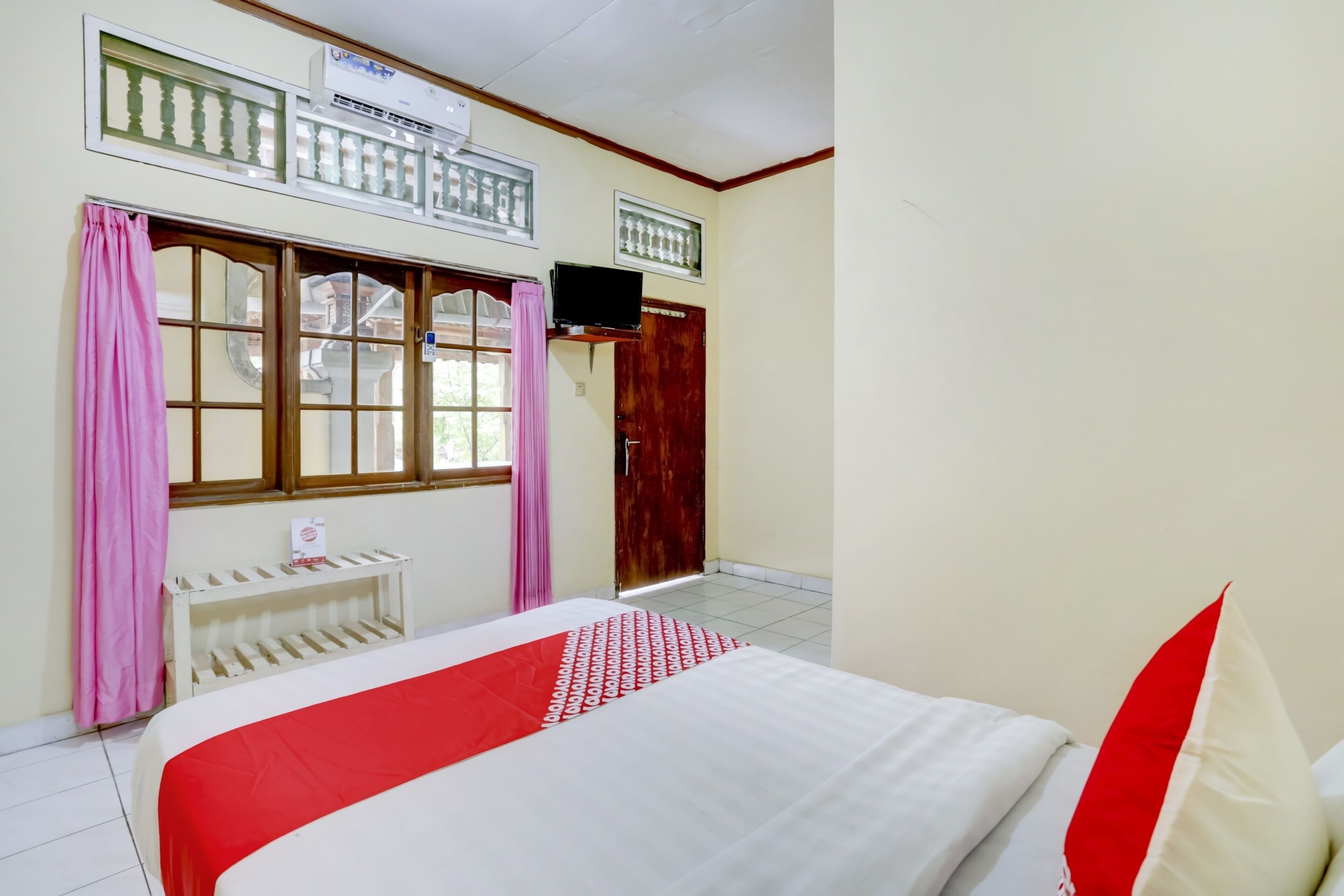 Un dormitorio con cama, televisor montado en la pared, ventana con cortinas y puerta.