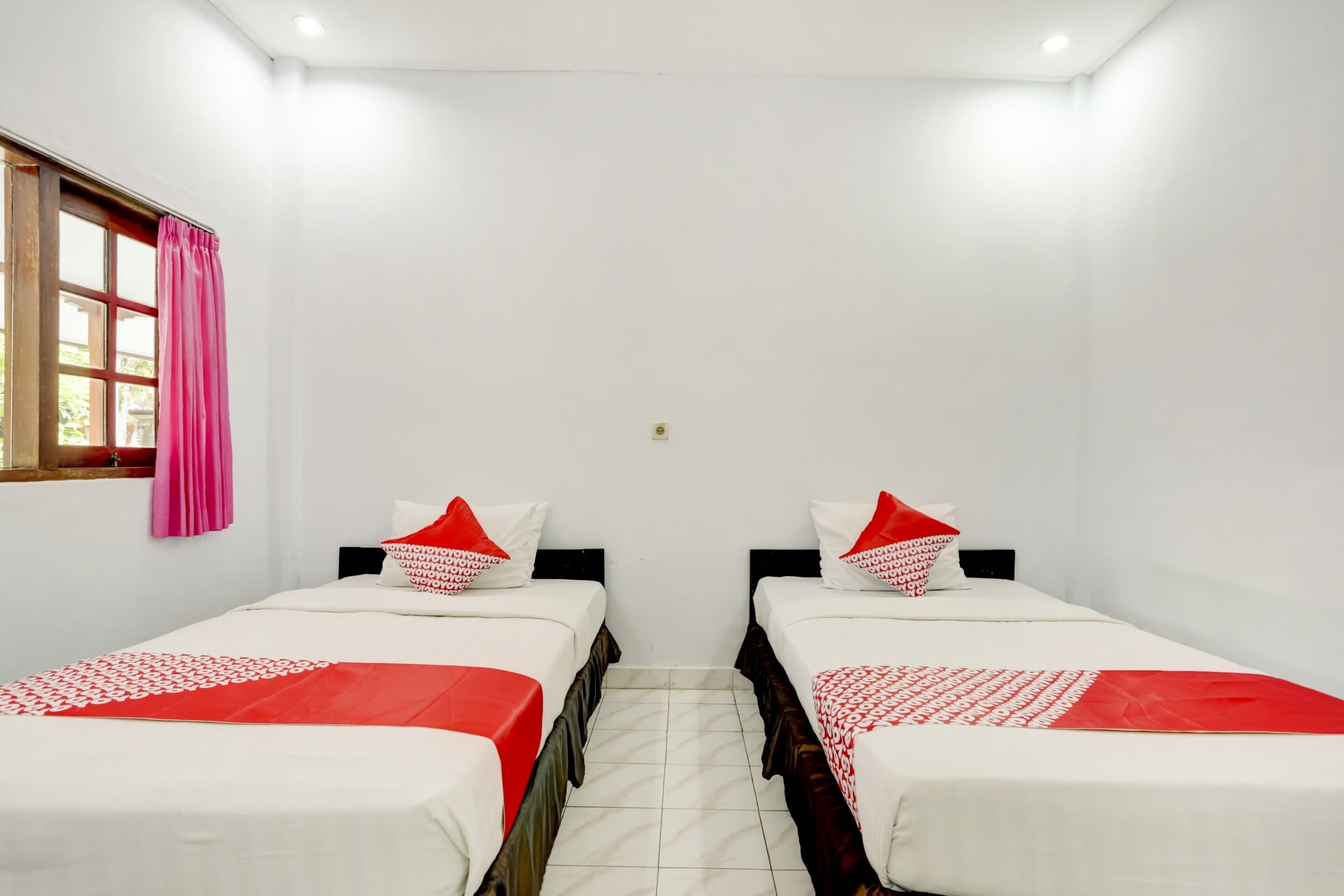 Quarto com duas camas, cada uma com colcha vermelha e branca, almofadas brancas e uma janela com cortinas cor de rosa.