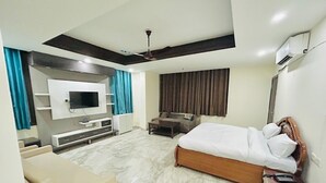 A modern hotel room with a large bed, a TV mounted on the wall, a ceiling fan, and a seating area with a sofa and coffee table. - Roomshala 074 Hotel Royal Inn (Noida)
