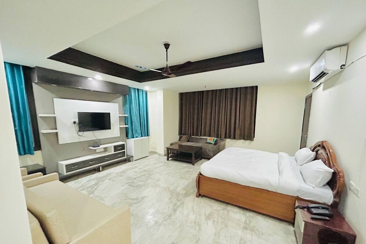 A modern hotel room with a large bed, a TV mounted on the wall, a ceiling fan, and a seating area with a sofa and coffee table.