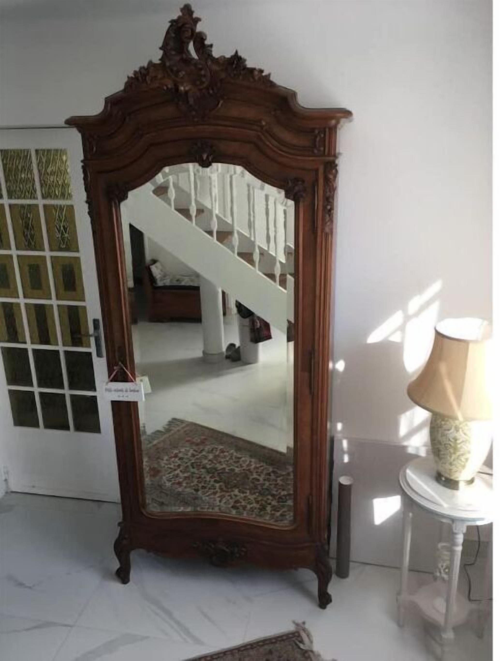 Ein wandmontierter Spiegel mit Holzrahmen und dekorativer Oberseite, platziert in einem Zimmer mit Treppe und einer Lampe auf einem kleinen Tisch.