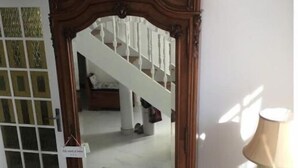 A wooden-framed mirror with a decorative top, placed in a room with a staircase and a lamp on a small table.