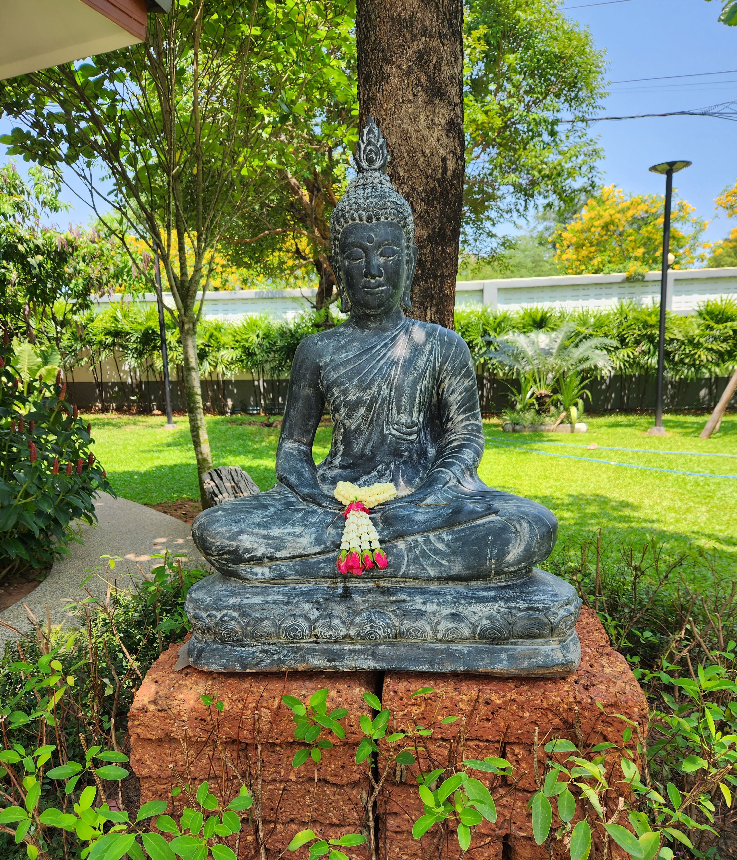 A seated Buddha statue with a garland, surrounded by greenery and trees.