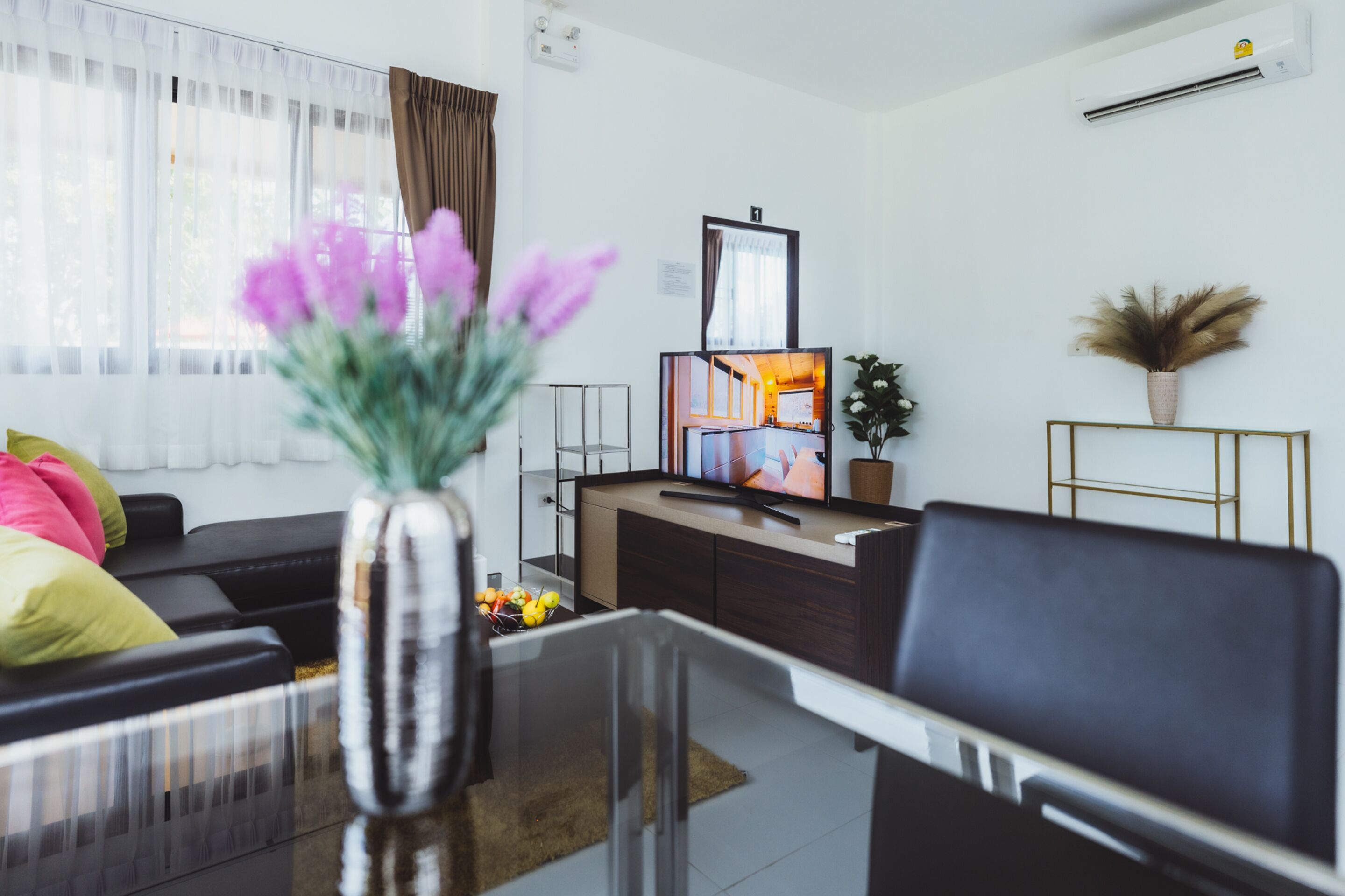 A modern living room with a sofa, a coffee table, and a TV. There is a window with curtains, a plant, and a vase with flowers.
