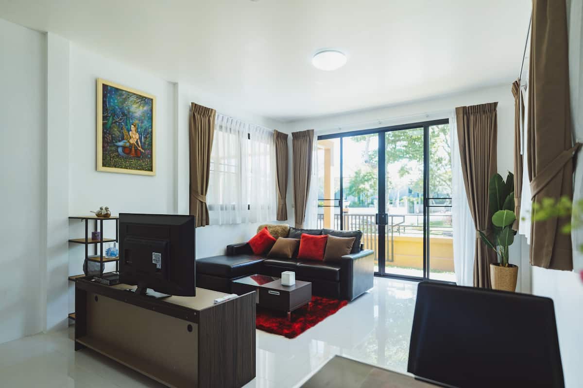 Een moderne woonkamer met een flatscreen televisie, een bank en een salontafel. Er is een groot raam met gordijnen, een balkon en een schilderij aan de muur.