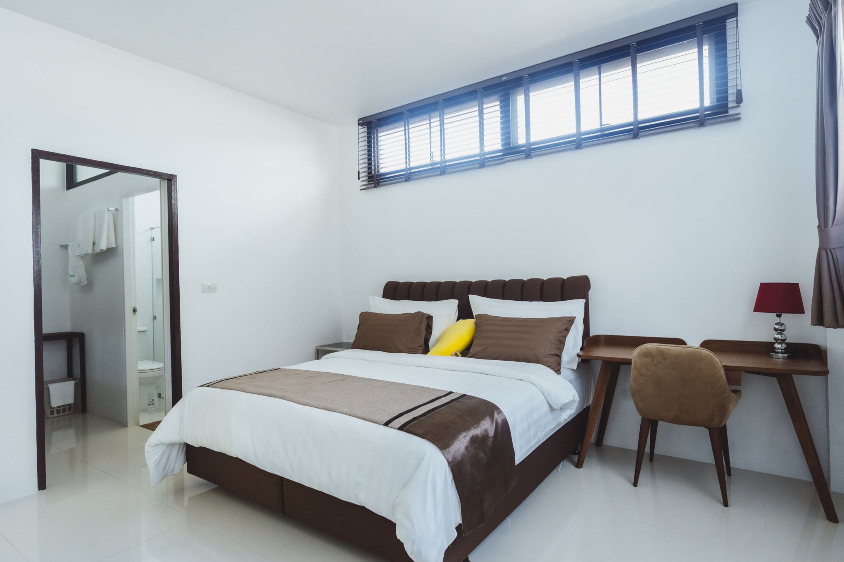 A modern bedroom with a large bed, a desk, and a chair. There is a window with blinds, a mirror, and a door leading to a bathroom.