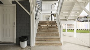 Un escalier en bois avec des rampes blanches mène à une zone abritée d’où l’on peut admirer une maison au loin.