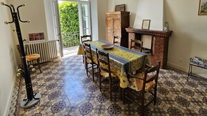 Un coin repas avec un sol à motifs, une table recouverte d’une nappe colorée, des chaises en bois, un radiateur, un porte-manteau, une cheminée et une bibliothèque.