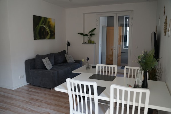 Un salon moderne avec un canapé gris foncé, des chaises blanches, une table blanche et une porte en verre donnant sur un balcon.