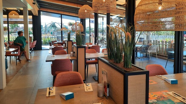 Een modern restaurant met hoge plafonds, zichtbare balken en grote ramen. Tafels staan er met oranje stoelen, en aan het plafond hangen decoratieve rieten lampen.
