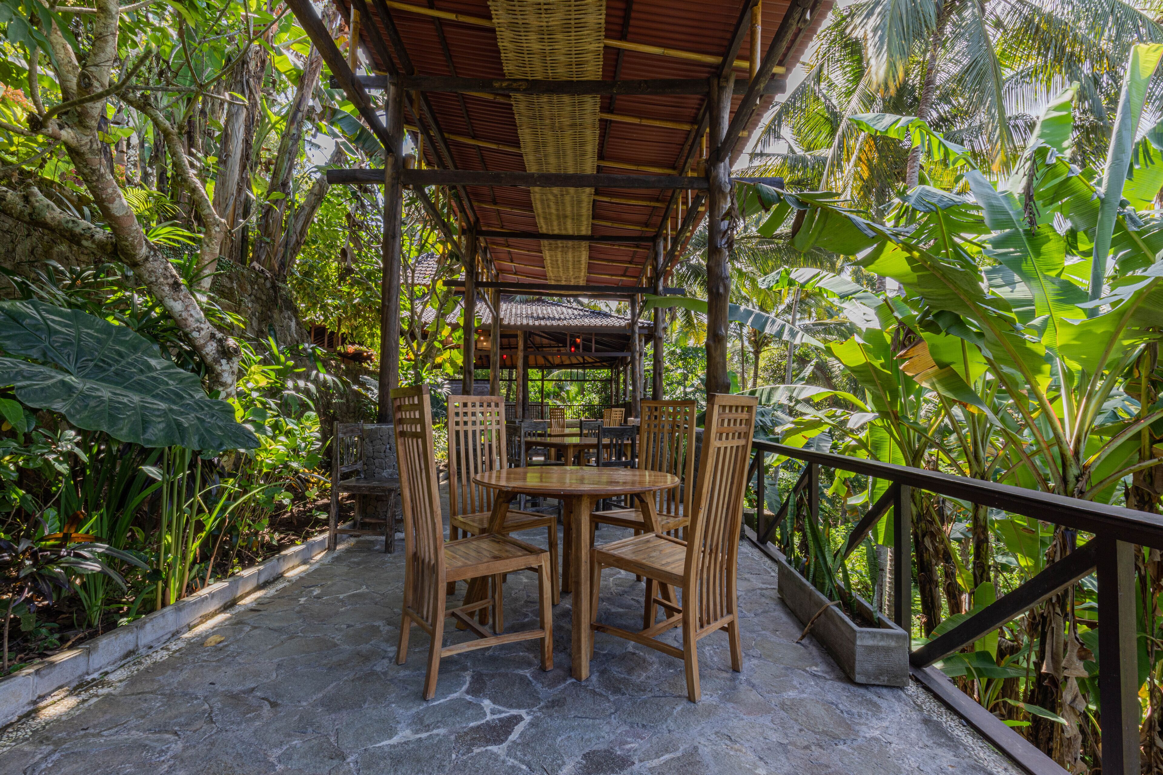 An outdoor wooden pavilion with a tiled roof, surrounded by lush tropical vegetation and stone flooring.
