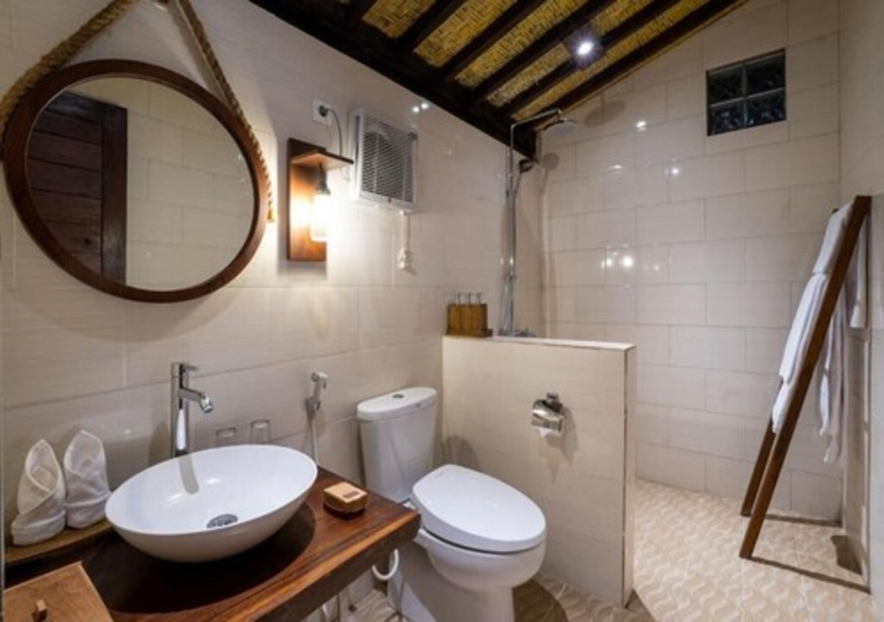 A bathroom with a wooden vanity, a round mirror, a white sink, a toilet, and a shower area.