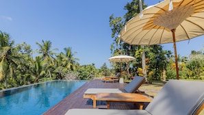 Eine Holzterrasse mit Liegestühlen und Sonnenschirmen mit Blick auf einen Swimmingpool, umgeben von üppiger Bepflanzung.