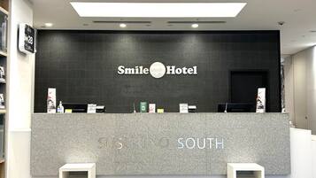 A reception desk at Smile Hotel with a dark wall and a sign that reads "Smile Hotel." There are two white benches in front of the desk, and various informational posters are displayed on the wall.