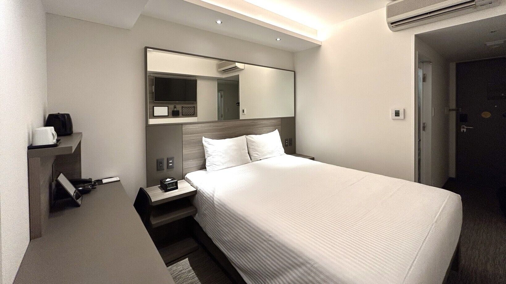 Una camera d'albergo con un letto, un comodino, uno specchio, una TV e un condizionatore d'aria.