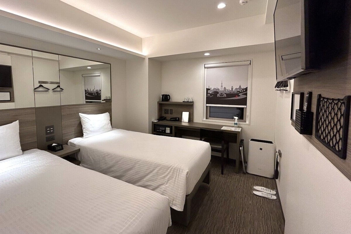 Ein Hotelzimmer mit zwei Betten, einem Schreibtisch, einem Spiegel, einem Fernseher und Blick auf die Stadt.