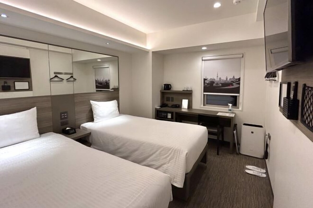 A hotel room with two beds, a desk with a chair, a small refrigerator, a mirror, and a window with a cityscape view.
